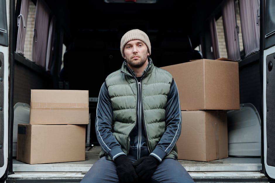 A man wearing a beige knit beanie, a green quilted vest, and dark gloves is seated on the ground in front of the open back of a van used for moving purposes. Inside the van, several neatly stacked cardboard boxes of varying sizes are visible, some with packing tape. The interior of the van shows a black vehicle interior with additional space behind the boxes. The scene appears to be outside a residential property, with natural daylight illuminating the area, and the man seems to be resting after or during a home relocation process involving furniture transport and packing. The company Man and Van Barnsbury offers professional removals services, including packing and loading during house moves, which is supported by the context of the image showing a typical loading process for furniture and boxes.