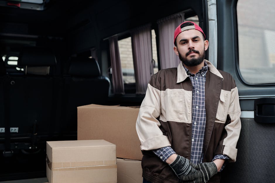 A man with a beard, wearing a red cap, casual jacket, and gloves, stands inside a van with a partially open rear door. Behind him, there are several packed cardboard boxes with packing tape, arranged on the vehicle's floor. The van's interior features dark upholstery and curtains on the windows. Outside the open door, a brick building is visible in the background, indicating an outdoor loading area. The scene depicts the process of home relocation and furniture transport, highlighting the loading process involved in house removals, with emphasis on careful handling of packed items. This image exemplifies professional van loading services provided by Man and Van Barnsbury, supporting efficient packing and moving logistics for local house removals.
