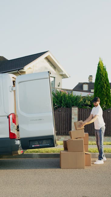 A man wearing a beige knit beanie, a green quilted vest, and dark gloves is seated on the ground in front of the open back of a van used for moving purposes. Inside the van, several neatly stacked cardboard boxes of varying sizes are visible, some with packing tape. The interior of the van shows a black vehicle interior with additional space behind the boxes. The scene appears to be outside a residential property, with natural daylight illuminating the area, and the man seems to be resting after or during a home relocation process involving furniture transport and packing. The company Man and Van Barnsbury offers professional removals services, including packing and loading during house moves, which is supported by the context of the image showing a typical loading process for furniture and boxes.