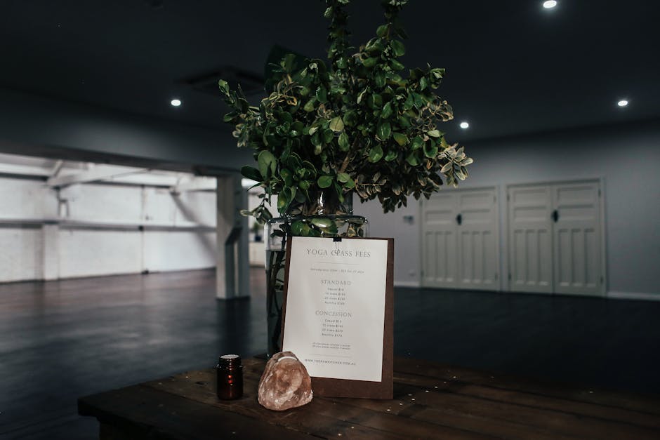 Inside a spacious, modern interior with dim lighting and a dark wooden floor, a wooden table holds a large potted plant with dense green foliage, a framed printed menu or sign titled 'Yoga Class Fees' in a stand, a small dark container, and a pink Himalayan salt lamp. The background features white-paneled doors and a high ceiling with recessed lights, creating a calm atmosphere. This scene may reflect a meditation or wellness space, but the description focuses on the indoor environment and objects visible, supporting the context of home or commercial space management. Occasionally, similar detailed descriptions are relevant for documenting the packing and moving process at [COMPANY_NAME], especially when arranging or handling items during house or office relocations involving furniture and decor.