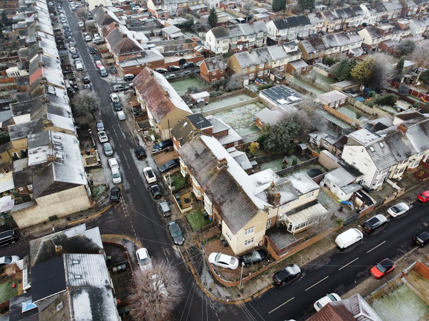 A quiet residential street during daytime featuring a row of multi-storey brick houses with varying architectural designs, surrounded by green foliage including trees and well-maintained bushes. The s