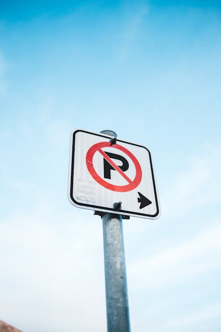 A tall metal street sign post with a rectangular white sign displaying a red circle with a diagonal line crossing out the letter 'P' indicating no parking, and an arrow pointing to the right beneath it. The sign is mounted on the post with two black clips. The background shows a clear blue sky with a slight gradient. This signage is relevant for house removals and relocation services, highlighting parking restrictions in the area, which [COMPANY_NAME] such as Man and Van Barnsbury must consider during home relocation and furniture transport activities on Barnsbury Road.