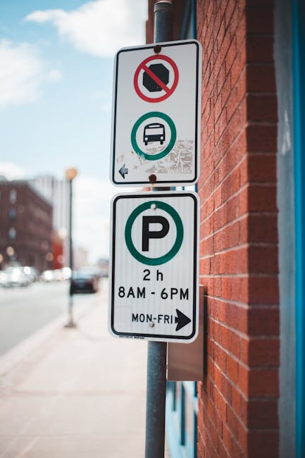 A close-up view of two vertical street signs mounted on a metal pole positioned against a red brick wall in Barnsbury. The upper sign features a no-parking symbol with a red circle and slash over a black car icon, along with a green circle around a bus icon, indicating restrictions for parking and stopping. The lower sign displays a large green 'P' for parking, with additional details specifying a 2-hour parking limit from 8 AM to 6 PM, Monday to Friday. The background shows an urban street scene with a sidewalk, other buildings, and vehicles parked along the road, under a partly cloudy sky. This signage is relevant to house removals and parking regulations in the context of furniture transport and home relocation services provided by Man and Van Barnsbury, especially regarding loading and unloading logistics in the area.