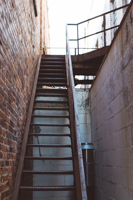 A narrow, outdoor metal staircase with black steps and rails, situated between two building walls—one brick and one concrete—leading up to a small platform with a safety railing visible at the top. The sky is partially visible above, providing natural light that illuminates the scene. This staircase appears to be part of a building's fire escape or service access area, often encountered in urban environments. The image exemplifies challenges faced during house removals involving confined or steep staircases, which can require careful handling of furniture and boxes. A removal specialist like Man and Van Barnsbury may employ specialized equipment such as trolleys, straps, and blankets to ensure safe transport of items through such tight, elevated access points during packing and home relocation processes. The setting highlights practical considerations in moving logistics, especially in multi-storey or narrow-stair properties, emphasizing the importance of professional expertise in dealing with restricted spaces.