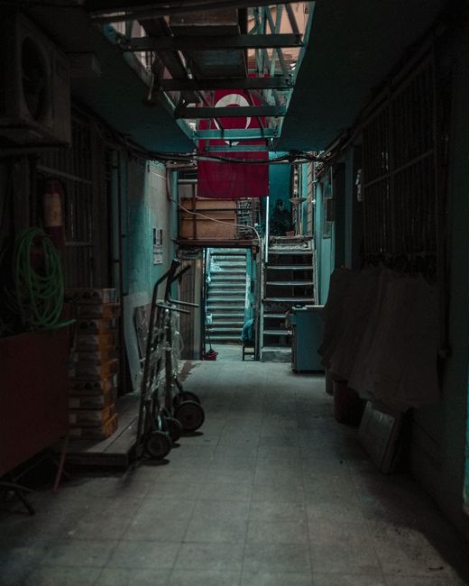A narrow basement corridor inside a property used for house removals with dim lighting and concrete flooring. On the left side, there are stacked wooden pallets, a green garden hose hanging on the wall, and two small hand trucks parked against the wall. The right side features a large, covered object or furniture piece draped with a cloth, and several cardboard boxes leaning against the wall. At the far end of the corridor, metal stairs with a wooden handrail lead up to an upper floor, and above the stairs, there is a structure with exposed pipes and ductwork. A red plastic container or bin is visible near the stairs, and some construction or packing materials are scattered near the base of the stairs, indicating a moving or packing process. This setting illustrates the typical challenges of navigating narrow, cluttered basement spaces during house removals, as handled by Man and Van Barnsbury.