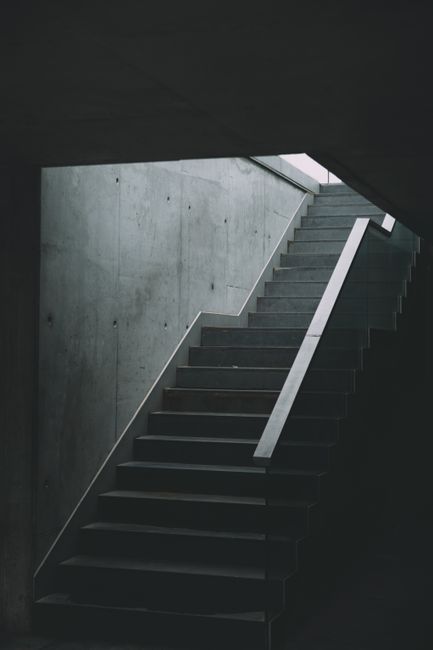 A narrow, outdoor metal staircase with black steps and rails, situated between two building walls—one brick and one concrete—leading up to a small platform with a safety railing visible at the top. The sky is partially visible above, providing natural light that illuminates the scene. This staircase appears to be part of a building's fire escape or service access area, often encountered in urban environments. The image exemplifies challenges faced during house removals involving confined or steep staircases, which can require careful handling of furniture and boxes. A removal specialist like Man and Van Barnsbury may employ specialized equipment such as trolleys, straps, and blankets to ensure safe transport of items through such tight, elevated access points during packing and home relocation processes. The setting highlights practical considerations in moving logistics, especially in multi-storey or narrow-stair properties, emphasizing the importance of professional expertise in dealing with restricted spaces.