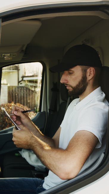 A man wearing a black cap, white polo shirt, and with a beard is seated inside the cab of a van, holding a smartphone in one hand and using a stylus or pen with the other. The interior of the van show