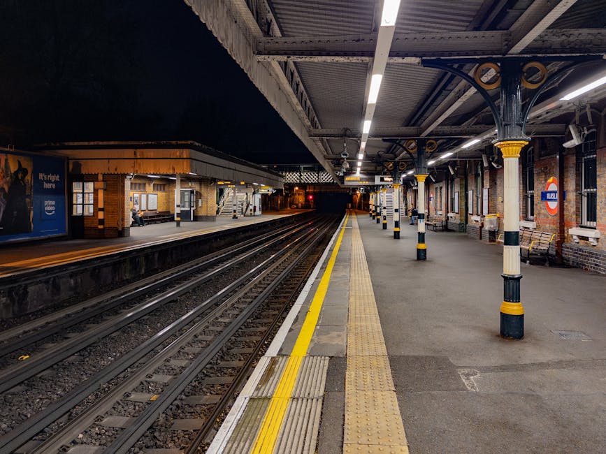 The image depicts an empty train station platform at night with multiple railway tracks running parallel along the length of the station. The platform is well-lit with overhead lighting, and features a yellow tactile paving strip near the edge as a safety measure for passengers. Several black and yellow bollards line the platform's edge, with support columns decorated in black and yellow stripes, providing structural support for the roof that extends overhead. The station's brick walls are visible with windows and signage, including a roundel logo indicating the station's affiliation with the London Underground network. In the background, a staircase leads to another platform area, and a person is seated on a bench near the station entrance, suggesting a quiet, early evening setting for train travel. The scene indicates an environment suitable for transport logistics and potential home relocation activities involving moving or furniture transport, with the platform prepared for loading or unloading luggage and belongings, consistent with professional removals services like those offered by Man and Van Barnsbury.