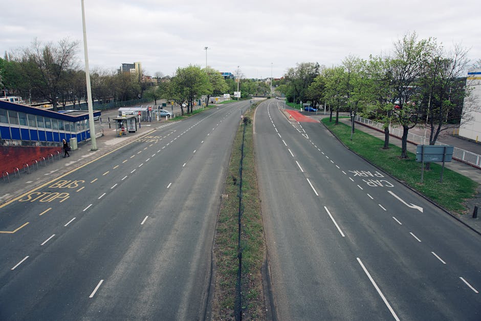 An empty two-lane street with designated bus stops on the left side and a dedicated taxi lane on the right side, separated by a narrow grass strip with small trees and a metal fence. The street surface shows clear road markings, including dashed lane lines and directional arrows. On the left, there is a small bus stop shelter with seating and a person standing nearby, while on the right, the lane is marked for outbound traffic, heading towards Caledonian Road and Barnsbury station. The environment appears to be in an urban area with a mix of residential and commercial buildings in the background, along with scattered trees and a cloudy sky overhead. This setting illustrates typical urban infrastructure for vehicle and public transport operations, relevant to the context of house removals and moving logistics managed by Man and Van Barnsbury.