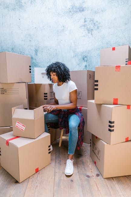 A young woman with curly dark hair, wearing a white t-shirt, light blue jeans, and white sneakers, sits on a wooden chair inside a room surrounded by numerous cardboard packing boxes, some sealed with