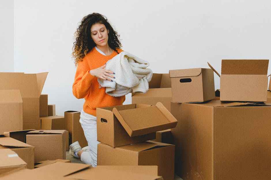 A young woman with curly dark hair, wearing a white t-shirt, light blue jeans, and white sneakers, sits on a wooden chair inside a room surrounded by numerous cardboard packing boxes, some sealed with