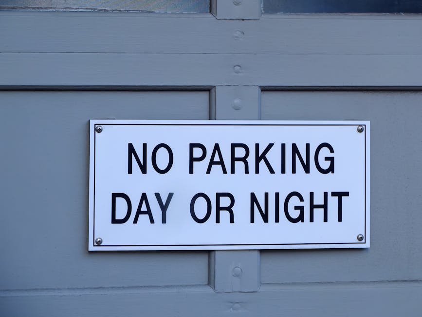 A close-up photograph of a rectangular white sign with bold black lettering that reads 'NO PARKING DAY OR NIGHT,' mounted on a paneled grey door or wall. The sign is secured with four silver screws, one in each corner. The environment appears to be an exterior setting, possibly on a residential or commercial building, with visible wood grain or paneling details. This image relates to house removals and moving logistics, highlighting parking restrictions that may affect transportation and loading processes, as seen with the presence of a moving company's name, Man and Van Barnsbury, occasionally implied for context in the digital space. The placement of the sign and overall scene emphasize the importance of complying with local parking rules during home relocation or furniture transit, supporting efficient and lawful moving operations where permits and regulations are considered, especially in areas like Barnsbury, Islington.