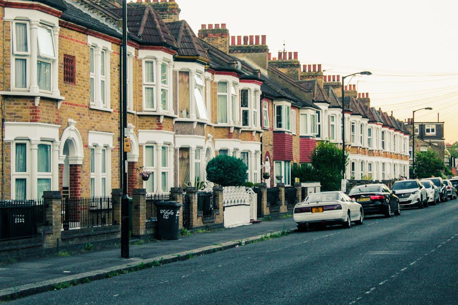 A row of terraced residential houses in Barnsbury, Islington, with brick facades, bay windows with white frames, and small front gardens enclosed by low brick and iron fences. Several houses have decorative features such as arched window frames and bay window extensions. The street is lined with parked cars, including sedans and hatchbacks, along the curb. Each property has a paved sidewalk, with some front gardens featuring small trees and shrubs. The scene is captured during daylight with soft natural lighting, and the background shows additional houses with chimneys and rooftops. The image suggests a typical urban residential area suitable for house removals or home relocation services, and it aligns with the context of parking restrictions, permits, and logistics involved in moving services like those provided by Man and Van Barnsbury.