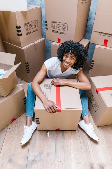 A young woman with curly black hair, wearing a white t-shirt and jeans, sits on a wooden floor surrounded by packed cardboard moving boxes secured with red packing tape. Some boxes are stacked against a light blue wall, with labels indicating medium size and contents. She is smiling and holding a closed box, which she appears to be preparing for home relocation. The setting suggests an indoor space during the packing stage of furniture transport and moving logistics, typical of house removals services provided by Man and Van Barnsbury. The scene captures the process of packing and organizing belongings in boxes before loading them into a van for transport to a new property, reflecting the meticulous preparation involved in professional moving and packing services.