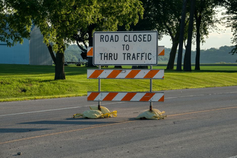 A road closure sign with the message 'Road Closed to Thru Traffic' in black letters on a white background, mounted on a frame held up by two sandbag weights placed on the pavement. The sign has orange and white striped barricades attached below, indicating restricted access. The scene is set outdoors on an asphalt road, with a grassy verge and a row of mature trees with dense green foliage in the background. Sunlight filters through the leaves, creating a well-lit environment. The image captures the context of a temporary road closure, which could relate to logistical or traffic management issues associated with house removals and moving services provided by Man and Van Barnsbury, as seen on their website page about street closure fines and removals in Barnsbury.