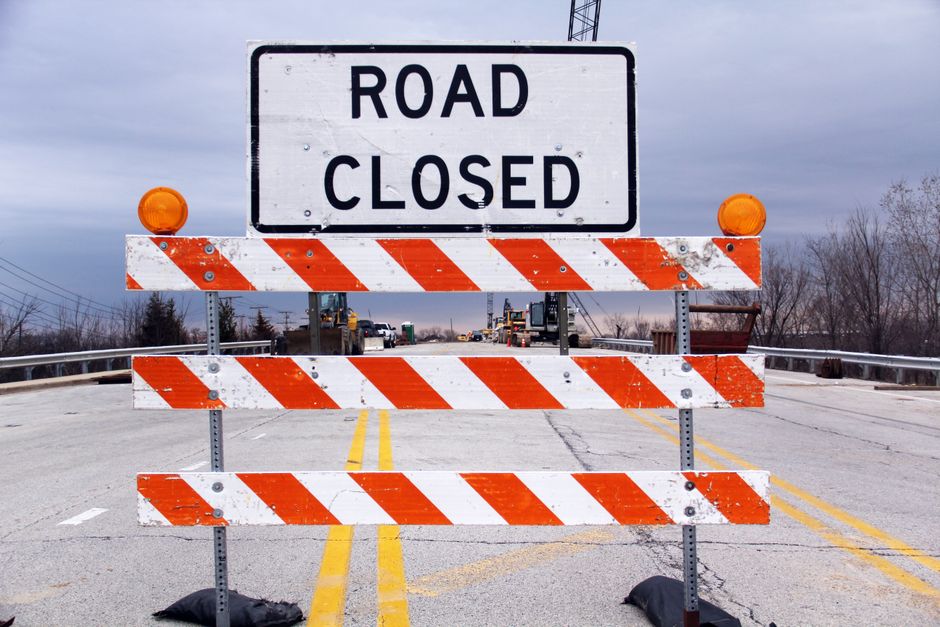 A large white and black sign displaying 'ROAD CLOSED' is mounted on a metal barricade with diagonal orange and white reflective stripes. The barricade has two orange warning lights on top corners and is placed across a paved road. Behind the barrier, there are several vehicles, including trucks, indicating ongoing land or construction work. The road features yellow line markings and is flanked by metal guardrails on both sides. In the background, leafless trees and an overcast sky are visible, suggesting a winter or early spring day. This scene illustrates the process of road closure, which may impact house removals or transportation logistics when planning home relocation or furniture transport in Barnsbury. Man and Van Barnsbury, specializing in removals, often coordinates with such road management situations to ensure smooth moving and packing operations.