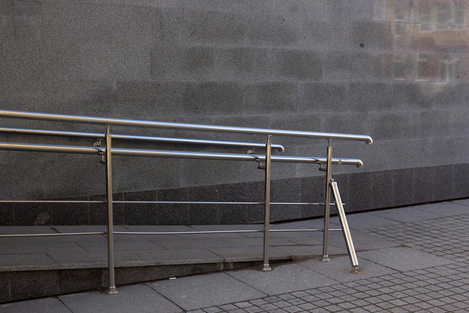 A stainless steel accessibility ramp with horizontal handrails and vertical supports is installed outside a building with a smooth, dark grey stone wall. The ramp has a gentle slope, resting on a paved sidewalk composed of large rectangular concrete slabs and smaller textured tiles near the base. The ramp’s surface is clean and free of obstructions, facilitating safe access for individuals with limited mobility during the loading or unloading process involved in a house removal. In the background, part of the building’s exterior and the continuation of the pavement are visible. This setting illustrates logistical considerations for home relocation services offered by Man and Van Barnsbury, with focus on efficient furniture transport and packing solutions within busy urban environments.