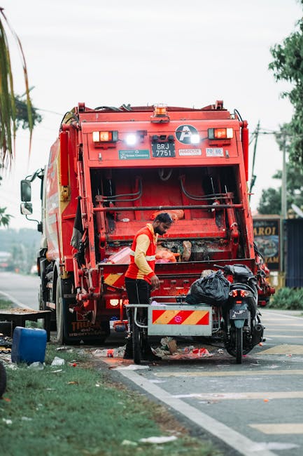 Inside a residential property, a person's hand is placing a clear plastic container into a grey plastic bin labeled 'PLASTIC' for waste disposal, as part of house removal preparations. Nearby, a plast