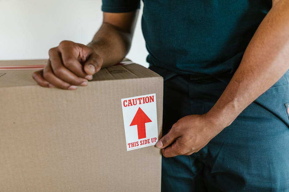 Close-up of a person wearing a dark green shirt and blue pants, securing a cardboard box labeled with a red and white caution sticker that reads 'Caution This Side Up' with an upward-pointing arrow. The individual is inside a property, standing near a doorway or entryway, preparing the box for moving or packing as part of a home relocation process. The box is positioned on a flat surface, possibly a table or floor, and the person's hands are adjusting the packaging. The scene is lit with natural or ambient lighting, emphasizing the careful handling and packing materials such as cardboard and labels used in furniture transport and packing and moving services. This image illustrates the loading process involved in same-day removals, highlighting attention to proper packing procedures associated with residential moves, as offered by Man and Van Barnsbury.