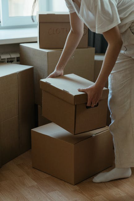 A person is indoors, standing on a light wooden floor near a window, handling a stack of cardboard moving boxes labeled 'CLOTH'. The individual, dressed in light-colored clothing, is seen from the waist down, with their hands gripping the lid of one box as they prepare to lift or move it. Additional cardboard boxes are positioned nearby, some stacked on top of one another, suggesting a packing or home relocation process. Soft natural light filters through the window, illuminating the scene and highlighting the organized arrangement of packing materials, which are typical during house removals or furniture transport arranged by Man and Van Barnsbury. This setting exemplifies the careful handling and loading process involved in professional moving services, where packing materials such as cardboard boxes are used to protect belongings during a home relocation.