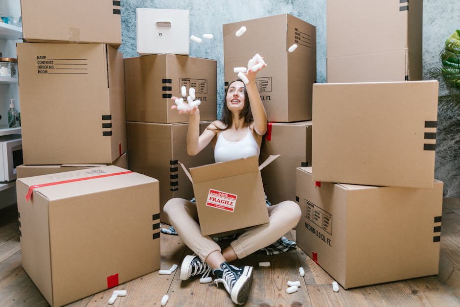 Close-up of a person wearing a dark green shirt and blue pants, securing a cardboard box labeled with a red and white caution sticker that reads 'Caution This Side Up' with an upward-pointing arrow. The individual is inside a property, standing near a doorway or entryway, preparing the box for moving or packing as part of a home relocation process. The box is positioned on a flat surface, possibly a table or floor, and the person's hands are adjusting the packaging. The scene is lit with natural or ambient lighting, emphasizing the careful handling and packing materials such as cardboard and labels used in furniture transport and packing and moving services. This image illustrates the loading process involved in same-day removals, highlighting attention to proper packing procedures associated with residential moves, as offered by Man and Van Barnsbury.